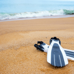 White and grey scuba diving fins on a white sand beach with the waves in the background